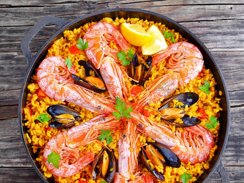 delicious seafood valencia paella with king prawns, mussels on savory saffron rice with spices and lemon wedges in pan, on wooden table, traditional spanish cuisine, view from above, close-up