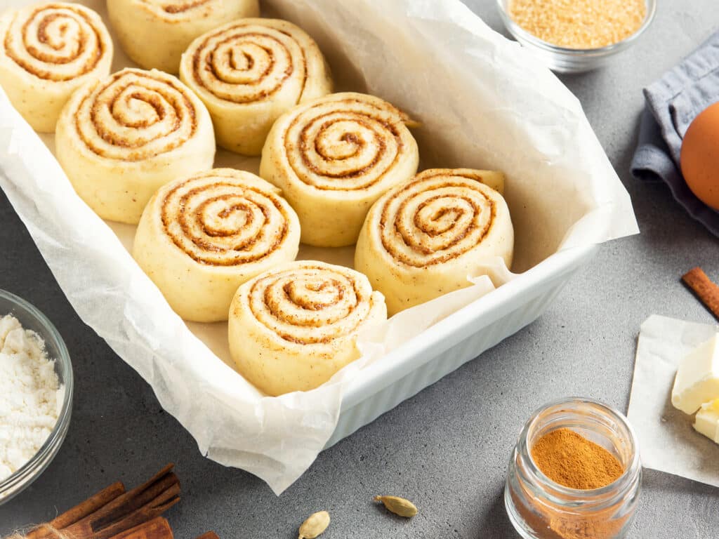 Cinnamon rolls or cinnabon, homemade recipe raw dough preparation sweet traditional dessert buns with pastry ingredients food on baking tray on grey background.