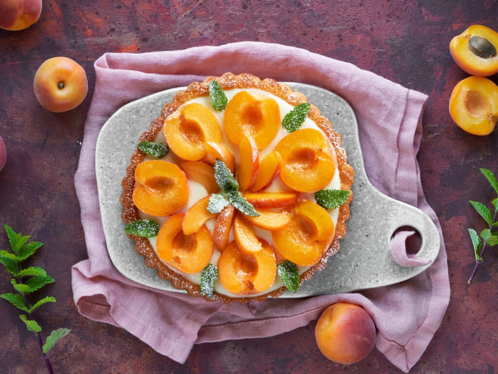 Top view of apricot tart with mint leaf on ceramic board with linen towel on dark red textured background
